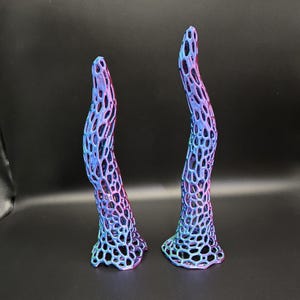 May include: Two abstract sculptures with a lattice design. The sculptures are tall and curved, with a gradient of colors from purple to blue to teal. The bases are flared, and the overall effect is organic and modern.