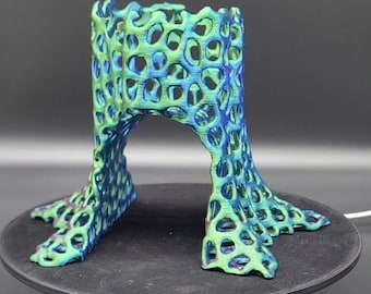 May include: A 3D printed sculpture with a green and blue iridescent finish. The sculpture is a hollow, open-ended cylinder with a lattice-like design. The cylinder is supported by three legs that extend out from the bottom.
