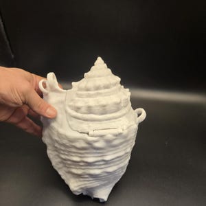 Könnte beinhalten: Ein weißer, muschelförmiger Behälter mit einem Klappdeckel und zwei kleinen Griffen. Der Behälter hat eine strukturierte Oberfläche und eine spiralförmige Oberseite, die einer Muschel ähnelt. Das Objekt befindet sich auf einem dunklen Hintergrund.