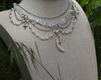 May include: A layered necklace with silver chains, white pearls, and iridescent beads. The necklace features a crescent moon pendant with a star and a star charm. The jewelry is displayed on a mannequin.