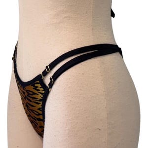 May include: A thong-style undergarment with a tiger stripe pattern in shades of brown and gold. The edges are trimmed with black fabric and gold-colored metal hardware. The straps are black.