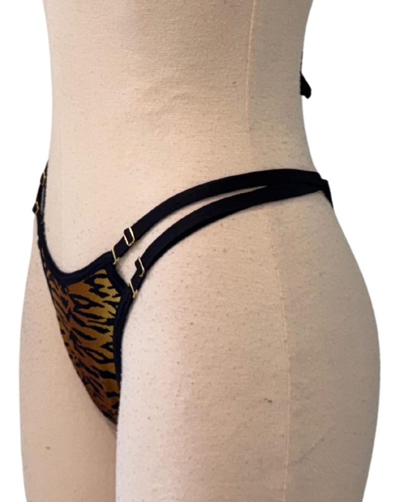 May include: A thong-style undergarment with a tiger stripe pattern in shades of brown and gold. The edges are trimmed with black fabric and gold-colored metal hardware. The straps are black.