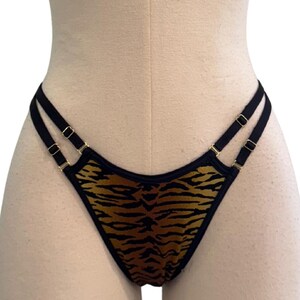 May include: A thong-style undergarment with a tiger stripe pattern in shades of brown and black. The straps are black with gold-colored hardware. The garment is displayed on a mannequin.