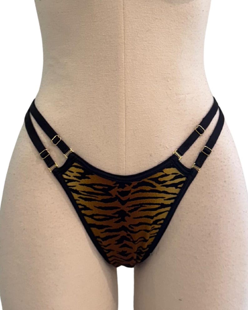 May include: A thong-style undergarment with a tiger stripe pattern in shades of brown and black. The straps are black with gold-colored hardware. The garment is displayed on a mannequin.