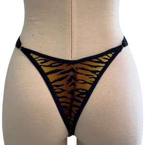 May include: A thong-style bottom with a tiger stripe pattern in shades of brown and black. The edges are trimmed with black fabric and gold-colored metal rings. The garment is displayed on a mannequin.