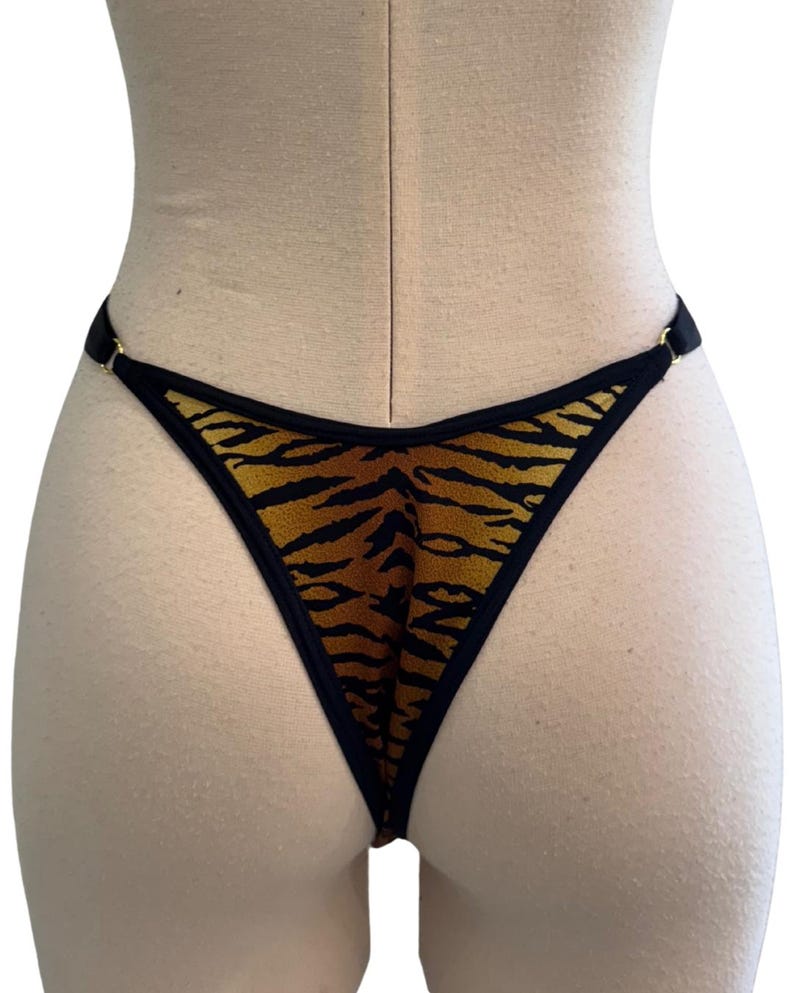 May include: A thong-style bottom with a tiger stripe pattern in shades of brown and black. The edges are trimmed with black fabric and gold-colored metal rings. The garment is displayed on a mannequin.