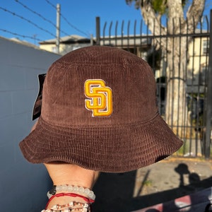 May include: A brown corduroy bucket hat featuring the San Diego Padres logo in gold and white. The hat has a classic bucket hat shape and is made of a ribbed corduroy fabric. The hat is a stylish accessory.
