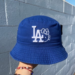 Puede incluir: Un sombrero de pescador azul con las letras bordadas en blanco "LA" y un diseño de Hello Kitty. El sombrero está hecho de una tela resistente y tiene una forma clásica de sombrero de pescador. El sombrero es de color azul vibrante.