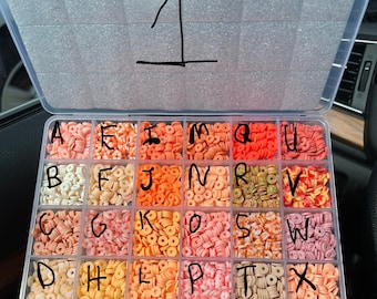 May include: A clear plastic storage box with 26 compartments, each filled with small, colorful, donut-shaped beads. The compartments are labeled with the letters of the alphabet, A through X.