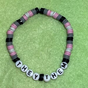Könnte beinhalten: Ein Armband aus schwarzen, grauen und rosa Perlen mit den Worten "THEY THEM" in weißen Perlen.