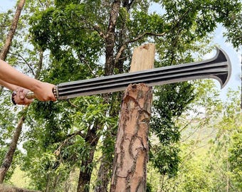 May include: A large, silver-colored sword with a curved blade and a ridged handle. The sword is held in front of a tree stump, with a forest background. The blade has a series of parallel grooves.
