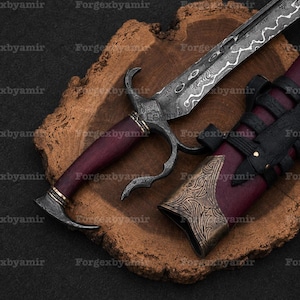 Puede incluir: Una daga con hoja de acero Damasco estampada, mango de color púrpura oscuro y guarda de metal curvada. La funda a juego es de color púrpura oscuro con adornos metálicos en color bronce y correas de cuero negro, sobre una rodaja de madera.