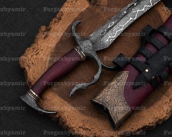 Puede incluir: Una daga con hoja de acero Damasco estampada, mango de color púrpura oscuro y guarda de metal curvada. La funda a juego es de color púrpura oscuro con adornos metálicos en color bronce y correas de cuero negro, sobre una rodaja de madera.