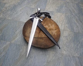 May include: A silver-toned steel dagger with a pointed blade and a crossguard, featuring a black handle. The dagger is accompanied by a black leather scabbard with integrated black leather straps and a metal buckle for secure attachment.
