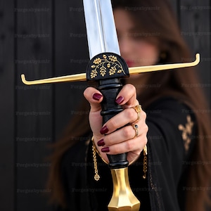 May include: A double-edged steel sword featuring a black grip, a gold-colored pommel, and a crossguard decorated with gold-colored oak leaf and acorn engravings. The crossguard ends in upward-curving tips, and the blade has a central fuller.