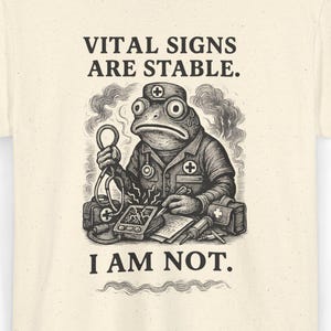 May include: Cream-coloured t-shirt with a detailed illustration of a frog in medical attire. The frog is surrounded by medical equipment, and the text "VITAL SIGNS ARE STABLE. I AM NOT." is printed above and below the frog.