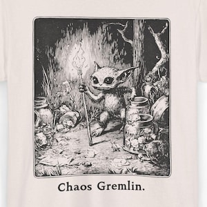 Può includere: Una maglietta beige con un'illustrazione in bianco e nero di una creatura che tiene un bastone in un ambiente boschivo. Il testo "Chaos Gremlin." è stampato sotto l'illustrazione.