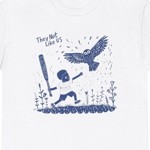 May include: White t-shirt with a blue illustration of a boy running with a bat and an owl flying above him. The text "They Not Like Us" is printed on the shirt.