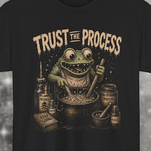 Puede incluir: Camiseta negra con una ilustración de una rana removiendo un caldero. El texto "TRUST THE PROCESS" está arriba. Frascos con las palabras "Paciencia", "Consistencia" y "Enfoque" rodean el caldero.