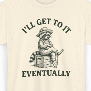 Puede incluir: Camiseta color crema con una ilustración relajada de un mapache con un overol y un sombrero, sentado en una caja de madera. El texto de arriba dice "I'll Get To It" y abajo, "Eventually."