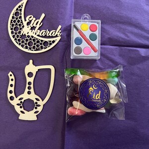 May include: A craft kit for children to celebrate Eid. The kit includes a wooden crescent moon cutout, a wooden teapot cutout, a small plastic container of watercolours and a brush, and a bag of colourful sweets with a sticker that says "Eid Mubarak".