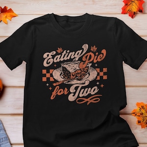 以下が含まれることがあります： 「Eating Pie for Two」と書いてあるヴィンテージスタイルのグラフィックがプリントされた黒いTシャツ。プレートの上にパイとフォークが描かれている。