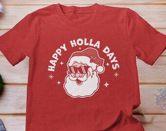 May include: Red t-shirt with a white graphic of Santa Claus wearing sunglasses and the text "HAPPY HOLLA DAYS". The shirt is displayed on a wooden surface with Christmas decorations, including ornaments and wrapped gifts.