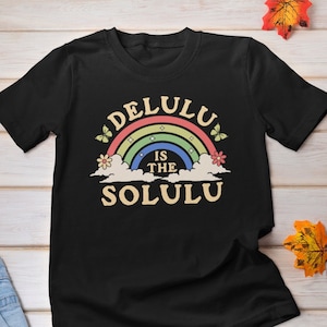 以下が含まれることがあります： レトロなフォントで「Delulu is the Solu」というテキストと虹が描かれた黒いTシャツです。