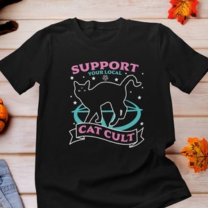 May include: Black t-shirt with a graphic of a black cat standing on a pentagram with stars and the text "Support Your Local Cat Cult".