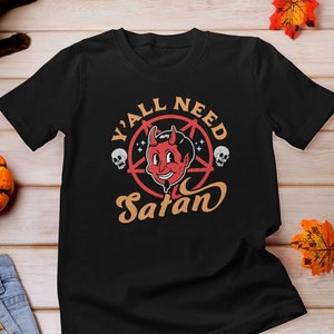 以下が含まれることがあります： オレンジ色の「Y'all Need Satan」の文字が入った五芒星をつけた赤い悪魔の漫画が描かれた黒いTシャツです。