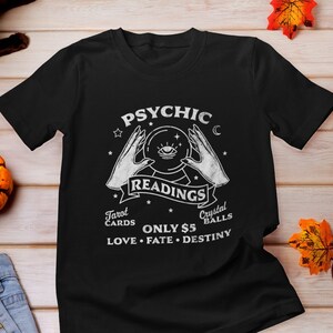 以下が含まれることがあります： 「Psychic Readings Tarot Cards Crystal Balls Only ¥5 Love Fate Destiny」という白い文字と、目をつけた水晶玉を2つの手で持っているグラフィックが描かれた黒いTシャツ。