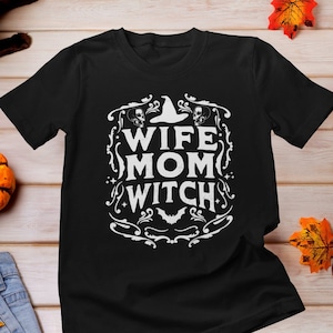 Pu&ograve; includere: Maglietta nera con un design grafico bianco che dice "Wife Mom Witch" con un cappello da strega, un pipistrello e teschi.