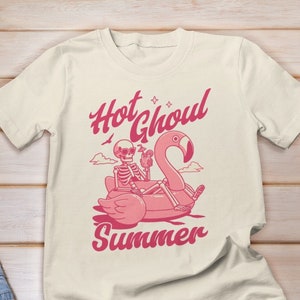 Może przedstawiać: Biały t-shirt z r&oacute;żowym nadrukiem przedstawiającym szkielet jadącego na dmuchanym flamingu z napisem "Hot Ghoul Summer".