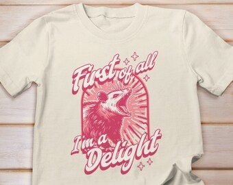 May include: A white t-shirt with a pink graphic of an opossum with the text "First of all I'm a Delight".