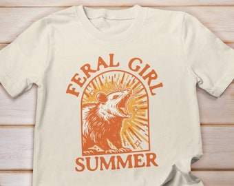 Op de afbeelding: Een wit T-shirt met een oranje afbeelding van een opossum met de tekst "Feral Girl Summer".