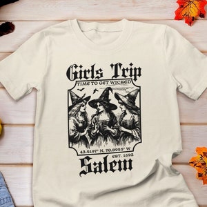 May include: A white t-shirt with a black graphic design of three witches with the text "Girls Trip Time to Get Wicked Salem EST. 1692" and coordinates 42.5197 N, 70.8993 W.