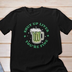 以下が含まれることがあります： 緑のクローバーのデザインと「Shut Up Liver You're Fine」というテキストが書かれた黒いTシャツ。