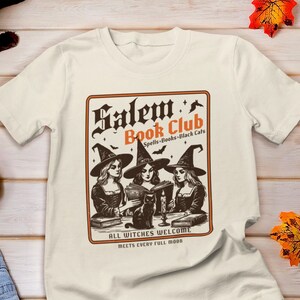 May include: A white t-shirt with a vintage-style graphic of three witches reading books in a book club. The graphic says "Salem's Book Club - Spells, Books, Black Cats" and "All Witches Welcome. Meets Every Full Moon."