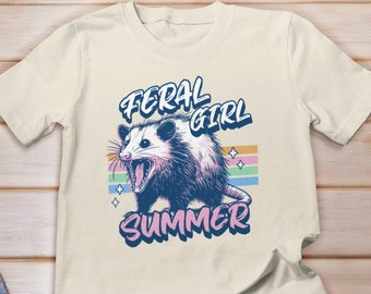 Könnte beinhalten: Ein beiges T-Shirt mit einem Opossum-Motiv und dem Text "Feral Girl Summer" in einem Regenbogen-Farbschema.