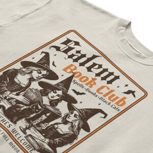 May include: A beige sweatshirt with a graphic print of three witches reading books in a room with a black cat. The graphic is in a brown and orange color scheme and says "Salem Book Club Spells, Books, Black Cats" and "All Witches Welcome Meets Every Full Moon".