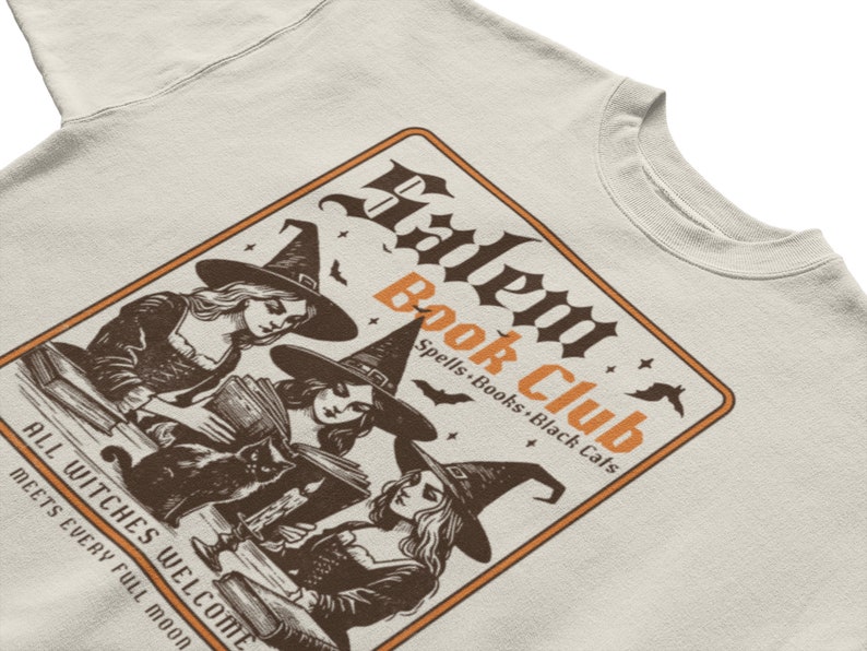 May include: A beige sweatshirt with a graphic print of three witches reading books in a room with a black cat. The graphic is in a brown and orange color scheme and says "Salem Book Club Spells, Books, Black Cats" and "All Witches Welcome Meets Every Full Moon".