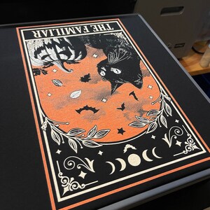 Pu&ograve; includere: Stampa grafica nera con un gatto nero, una zucca, pipistrelli e la scritta "THE FAMILIAR" su sfondo arancione. Il design &egrave; incorniciato da un bordo bianco ed elementi floreali decorativi, ideale per Halloween o decorazioni gotiche.