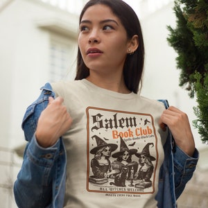 May include: A beige t-shirt with a vintage-style illustration of three witches reading books in a book club. The text reads "Salem Book Club Spells-Books-Black Cats" and "All Witches Welcome Meets Every Full Moon".