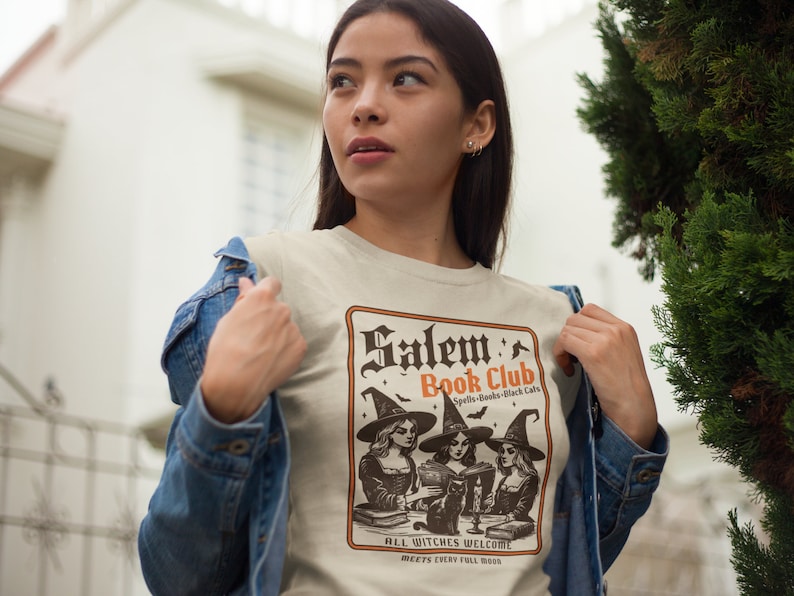 May include: A beige t-shirt with a vintage-style illustration of three witches reading books in a book club. The text reads "Salem Book Club Spells-Books-Black Cats" and "All Witches Welcome Meets Every Full Moon".