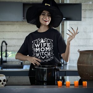 Pu&ograve; includere: Una donna con un cappello da strega nero e una maglietta nera con la scritta "Wife Mom Witch" sta mescolando una pentola in una cucina. Ha le unghie nere lunghe e sorride. Ci sono candele arancioni sul bancone di fronte a lei.