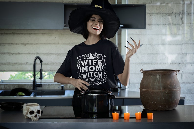 Pu&ograve; includere: Una donna con un cappello da strega nero e una maglietta nera con la scritta "Wife Mom Witch" sta mescolando una pentola in una cucina. Ha le unghie nere lunghe e sorride. Ci sono candele arancioni sul bancone di fronte a lei.