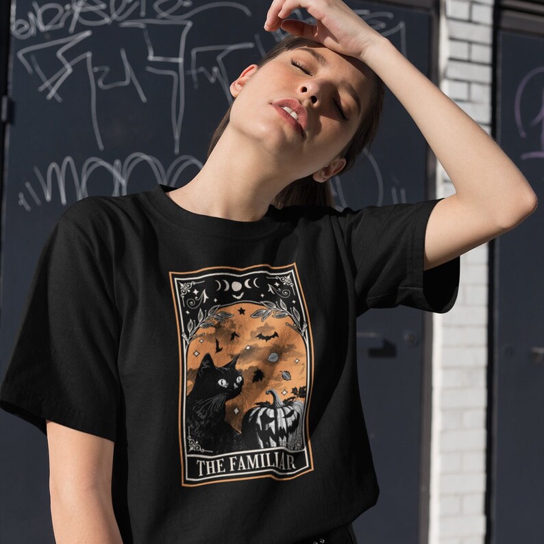 Pu&ograve; includere: T-shirt nero con un design a carta dei tarocchi. La grafica include un gatto nero, una zucca e pipistrelli su uno sfondo arancione. Il testo "THE FAMILIAR" &egrave; presente. La maglietta ha un girocollo.