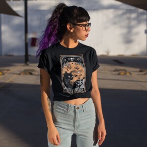 Pu&ograve; includere: T-shirt nera con una grafica di un gatto nero, zucca, pipistrelli e lune crescenti. Il testo "THE FAMILIAR" &egrave; sotto. La persona indossa occhiali e capelli viola.