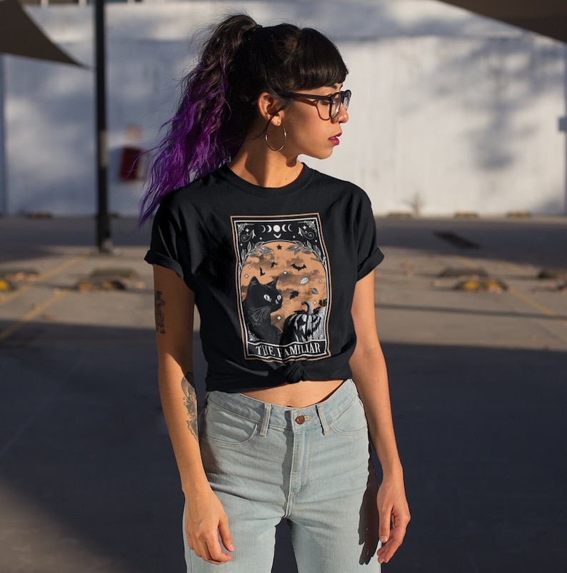 Pu&ograve; includere: T-shirt nera con una grafica di un gatto nero, zucca, pipistrelli e lune crescenti. Il testo "THE FAMILIAR" &egrave; sotto. La persona indossa occhiali e capelli viola.