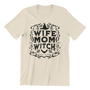 Pu&ograve; includere: Una maglietta beige con un design grafico nero che dice "Wife Mom Witch" con un cappello da strega e un pipistrello.
